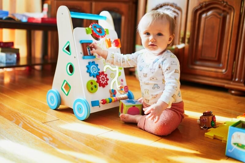 infant sitting on the floor playing with one hand on a push walker with an activity panel on the front