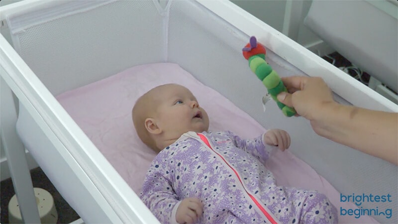 parent showing a baby a toy and baby watching that toy while lying in crib