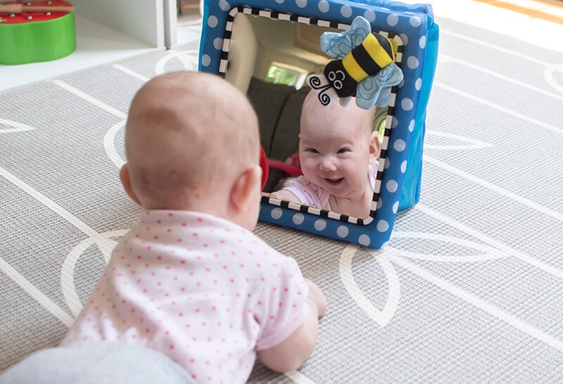 young baby in a pink shirt lying on the floor on her tummy smiling at her reflection in a soft mirror