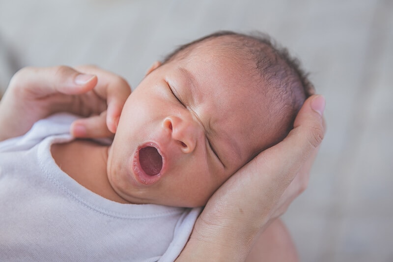 Young baby in parents' arms yawning, signalling the end of this round of awake time and now it's bed time.