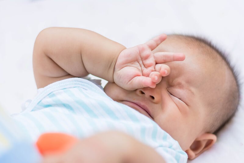 young baby sleeping in a swaddle with arms out, rubbing eyes