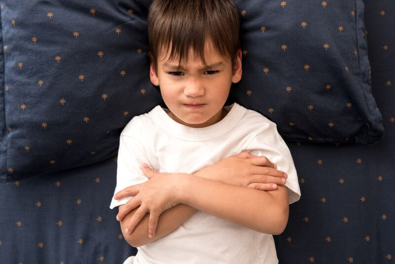 grumpy toddler who doesn't want to go to bed. Lying on bed, arms crossed, angry look on his face
