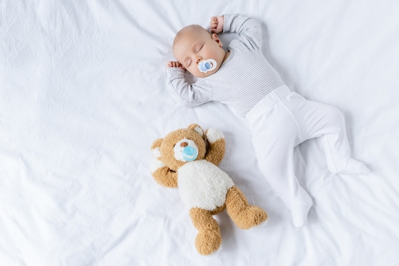 baby sleeping on her back in a crib with a teddy lying next to her