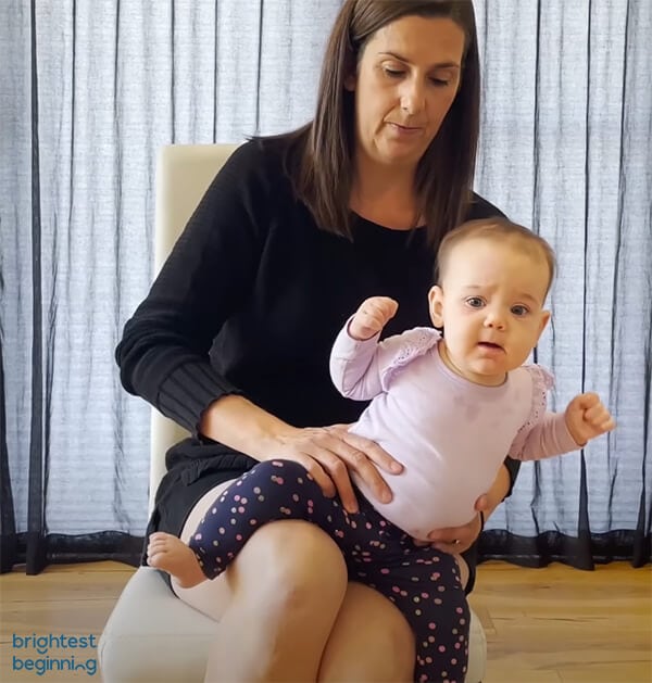 woman sitting on a white chair with a baby girl wearing a pink shirt sitting on her lap facing forwards. Woman gently tilts baby to the left while supporting her hips with her hands.
