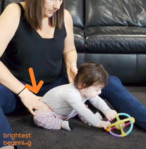 young child sat up in between woman's legs on the floor. He reaches forward to grab a coloured toy ball on the floor in front of him. Woman is pressing gently on his right hip to help him return to an upright position.
