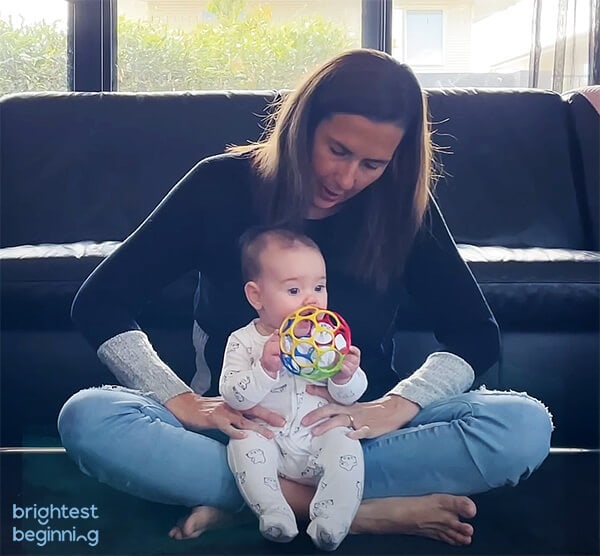 woman sitting with her legs crossed on the floor with a baby girl sitting in her lap facing outwards. Baby is holding a colourful toy ball